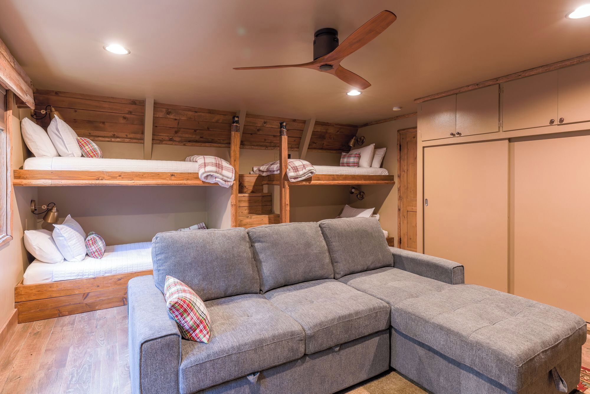 Una habitación con un sofá seccional gris, dos literas, paredes de madera, un ventilador de techo y un armario.