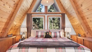 Een slaapkamer met een houten schuine kap, een groot bed met ruitdessin beddengoed, twee nachtlampjes en een raam met uitzicht op een bos.