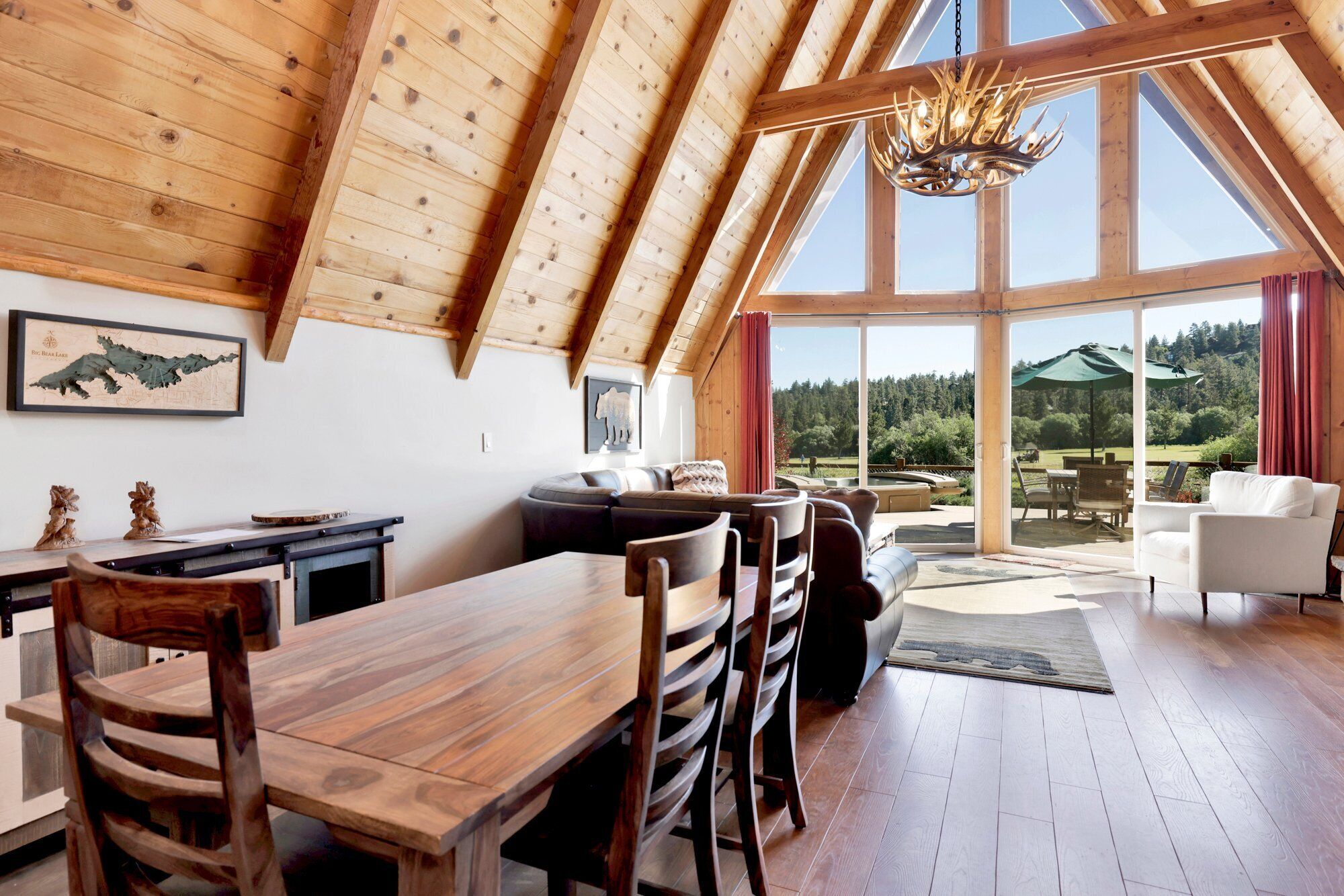 A spacious loft-style room with a high ceiling, wooden beams, a dining area with a long wooden table and chairs, a seating area with sofas, and a large window offering a view of a forested landscape.