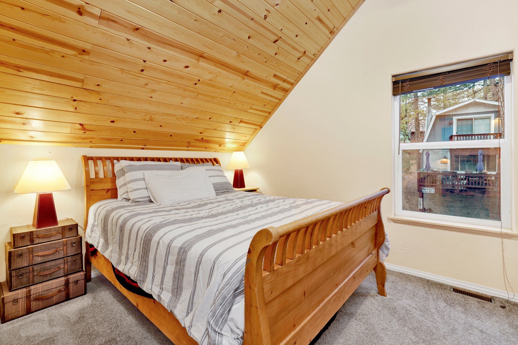 Quarto com teto de madeira, cama com roupa de cama listrada, duas luminárias de cabeceira e janela com vista para um edifício.