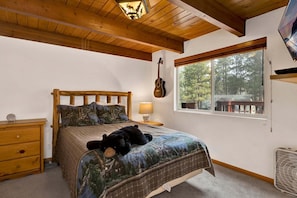 A bedroom with a wooden headboard, a bed with a forest-themed duvet, a guitar on the wall, a nightstand with a lamp, and a window with a view of trees.