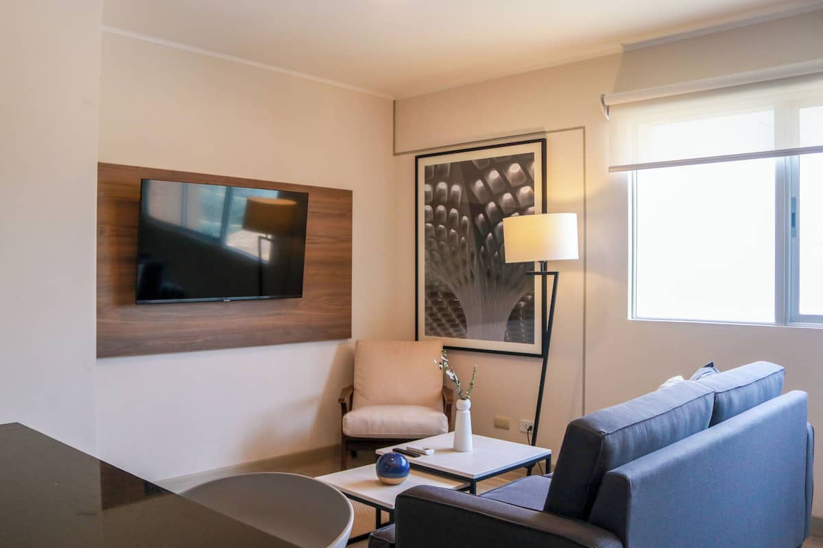 Quarto de hotel moderno com televisão de tela plana na parede, uma poltrona confortável, um sofá, uma mesinha com um vaso e um objeto decorativo azul, e uma janela grande que oferece luz natural.