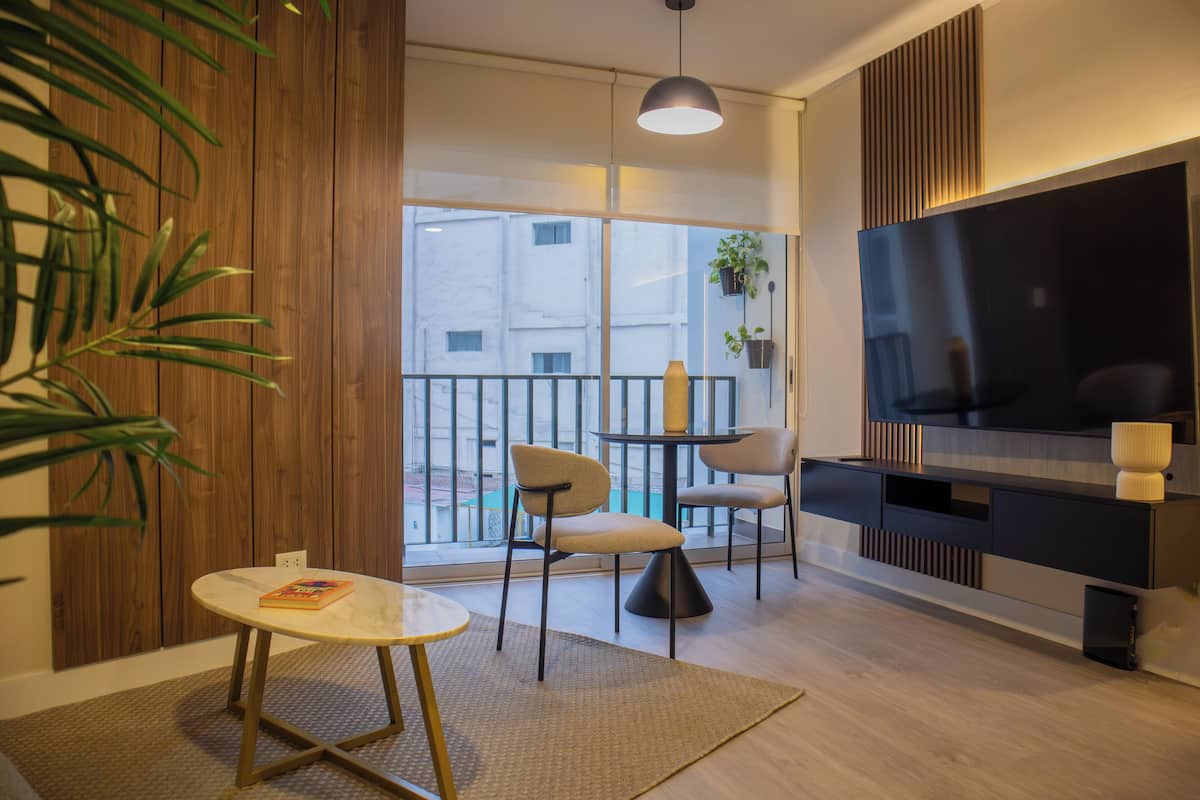 Uma sala de estar moderna com uma televisão, uma mesa redonda, duas cadeiras, um tapete pequeno e um vaso com planta.