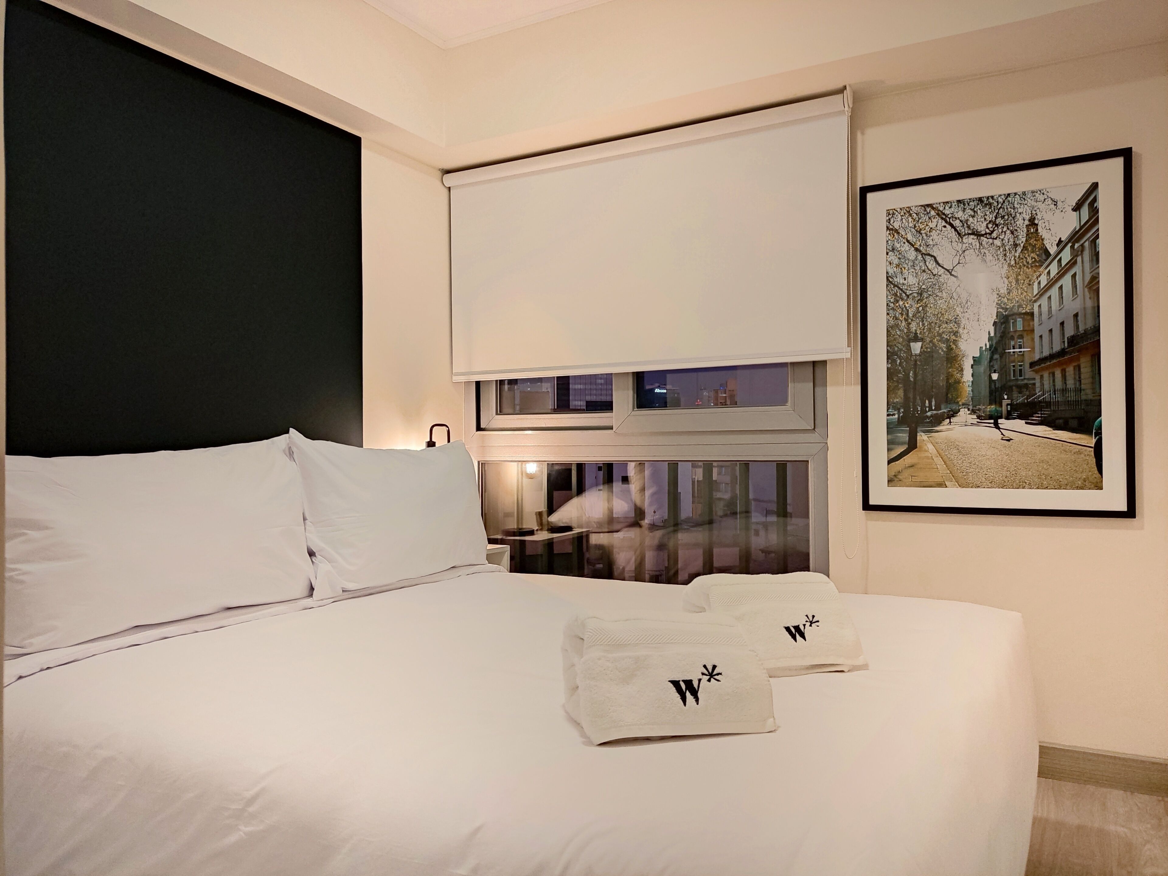 A hotel room with a large bed, a window with a view of a city street, and a framed picture on the wall.