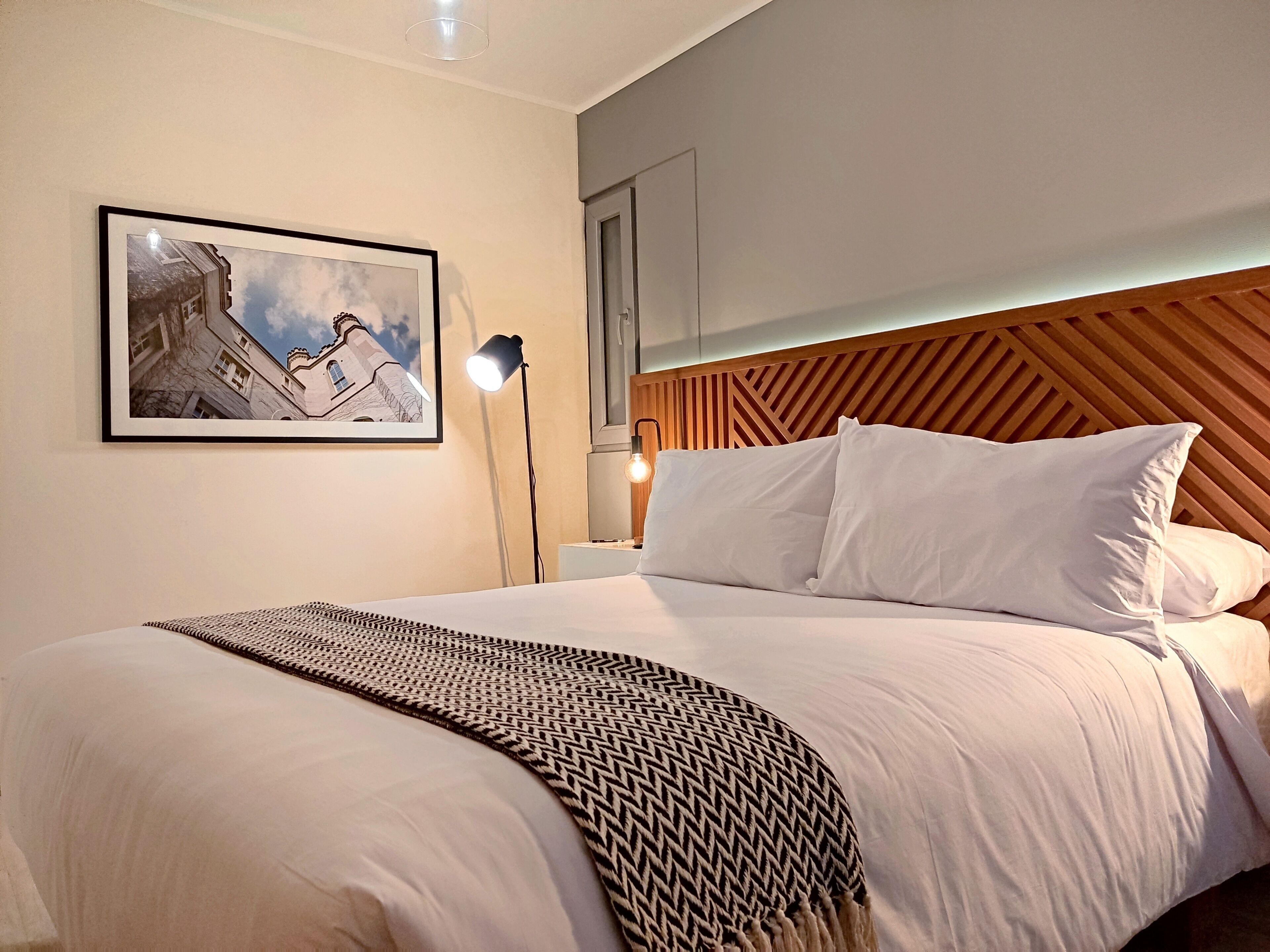 Un lit bien fait, avec du linge de lit blanc et un plaid à motifs, un tableau encadré au mur et une tête de lit moderne en bois.