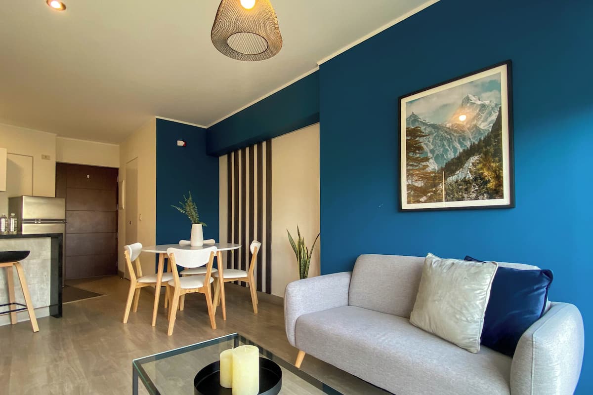 Uma sala de estar moderna com um sofá cinzento, uma mesa de jantar com cadeiras, uma área de cozinha e uma grande pintura de paisagem montanhosa na parede.