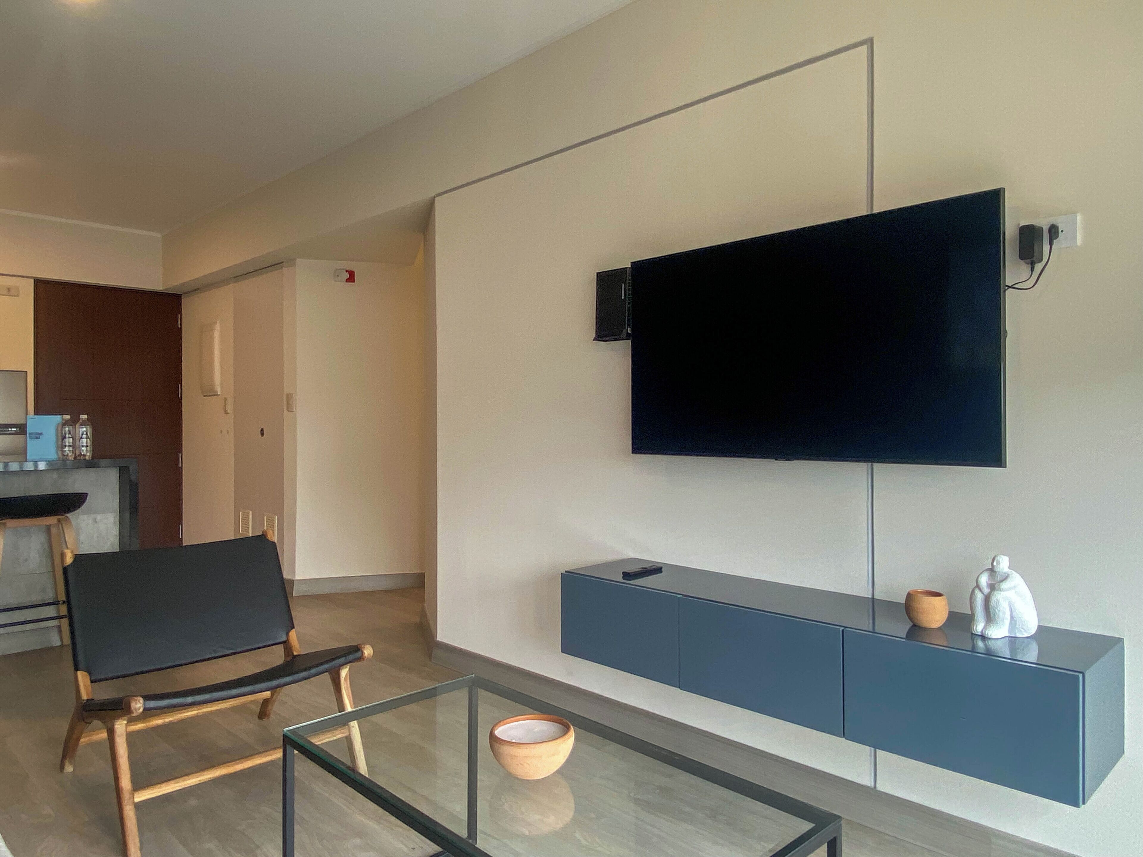 A modern living room with a flat-screen TV mounted on the wall, a glass coffee table, a black chair, and a blue console table with decorative items.