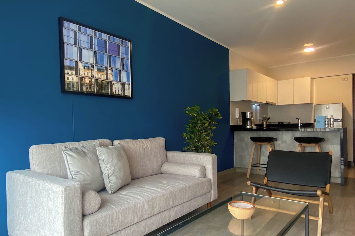 Uma sala de estar moderna com um sofá, uma mesa de centro de vidro e uma cozinha ao fundo.