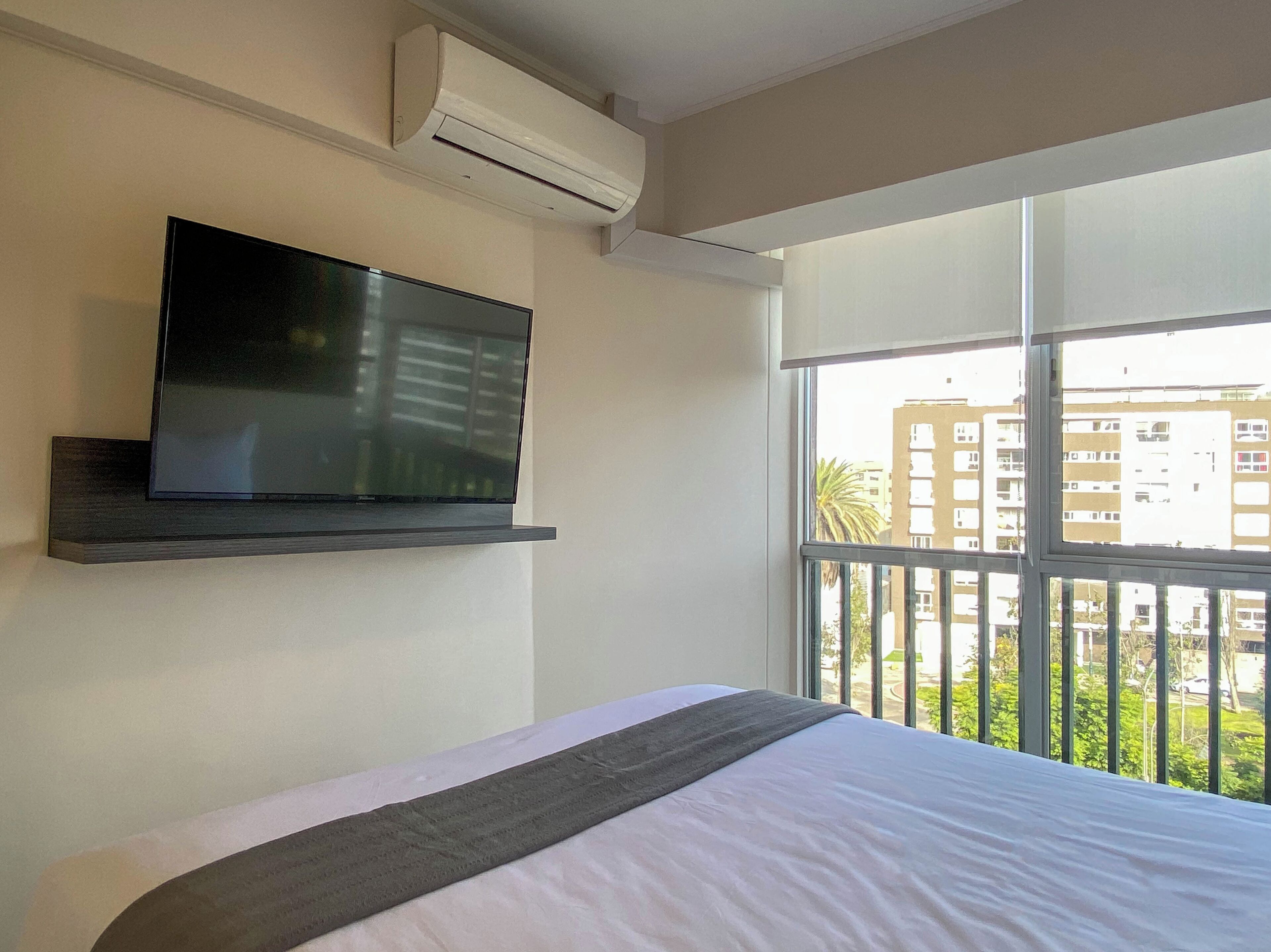 A hotel room with a bed, a mounted TV, an air conditioning unit, and a balcony with a view of buildings and greenery.