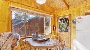 Interior de una cabaña de madera con una mesa de comedor, sillas, un refrigerador y una ventana, además de un extintor.