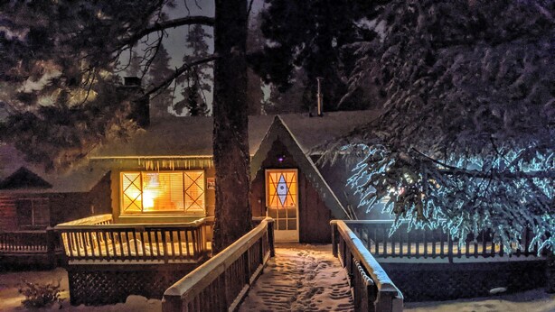 Un sendero cubierto de nieve que lleva a una cabaña de madera con puerta y ventanas de vidrio, rodeada de árboles.
