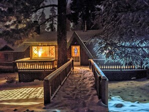 Un sendero cubierto de nieve que lleva a una cabaña de madera con puerta y ventanas de vidrio, rodeada de árboles.