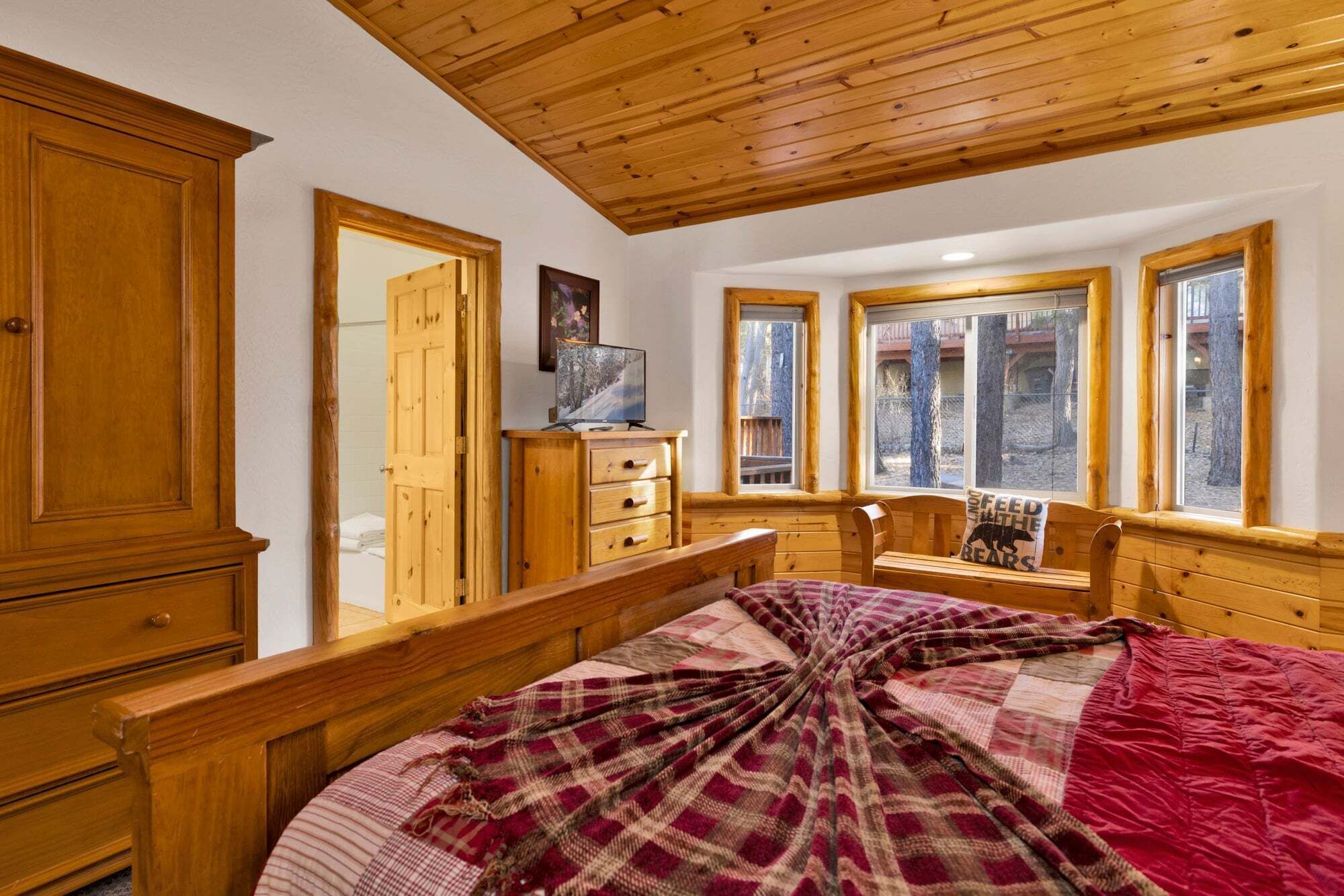 Un dormitorio con techo de madera, una cama grande con manta escocesa, un aparador de madera, una mesita de noche y una ventana con vistas a árboles.