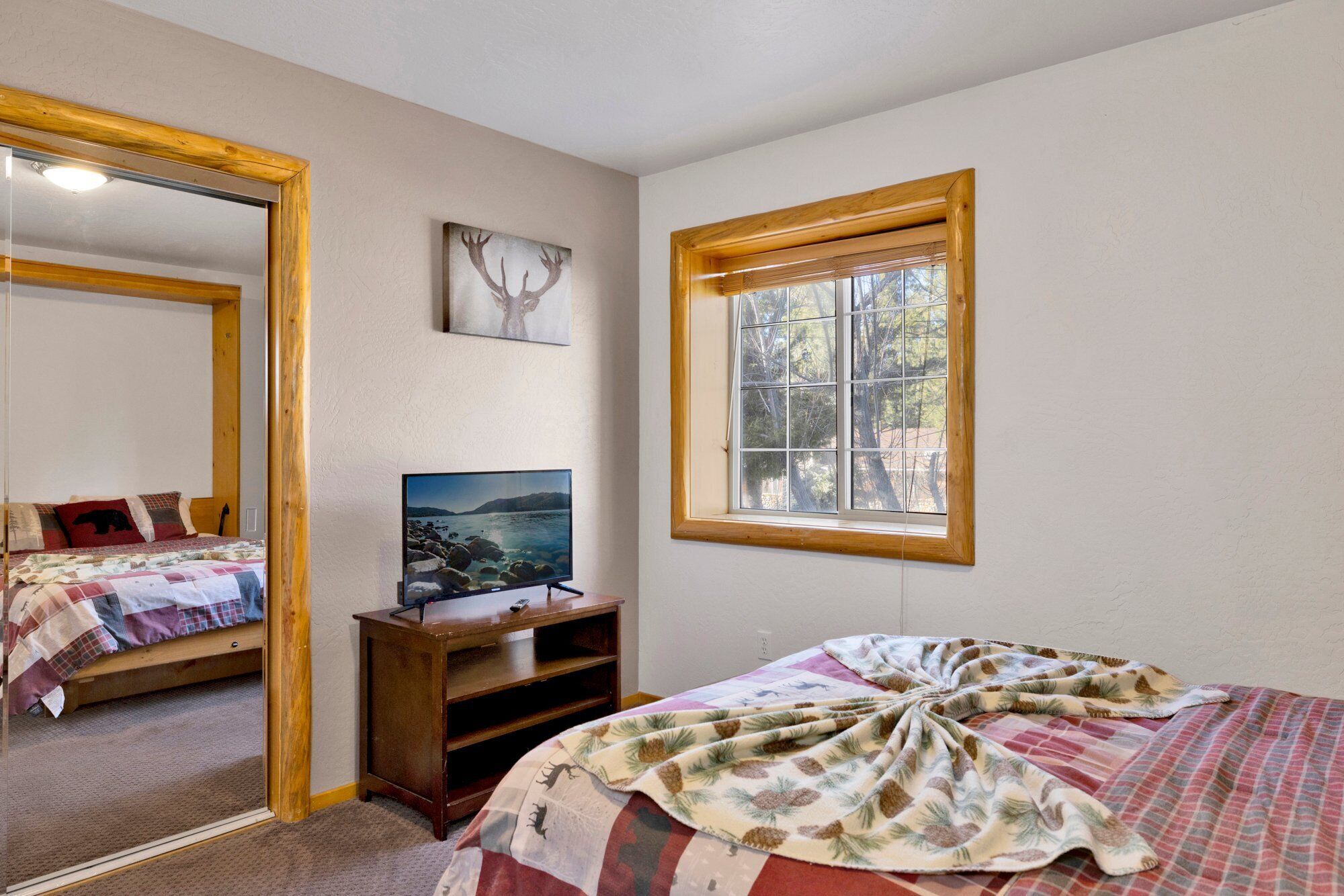 A bedroom with a bed, a TV on a wooden stand, a window with a view of trees, and a mirror.