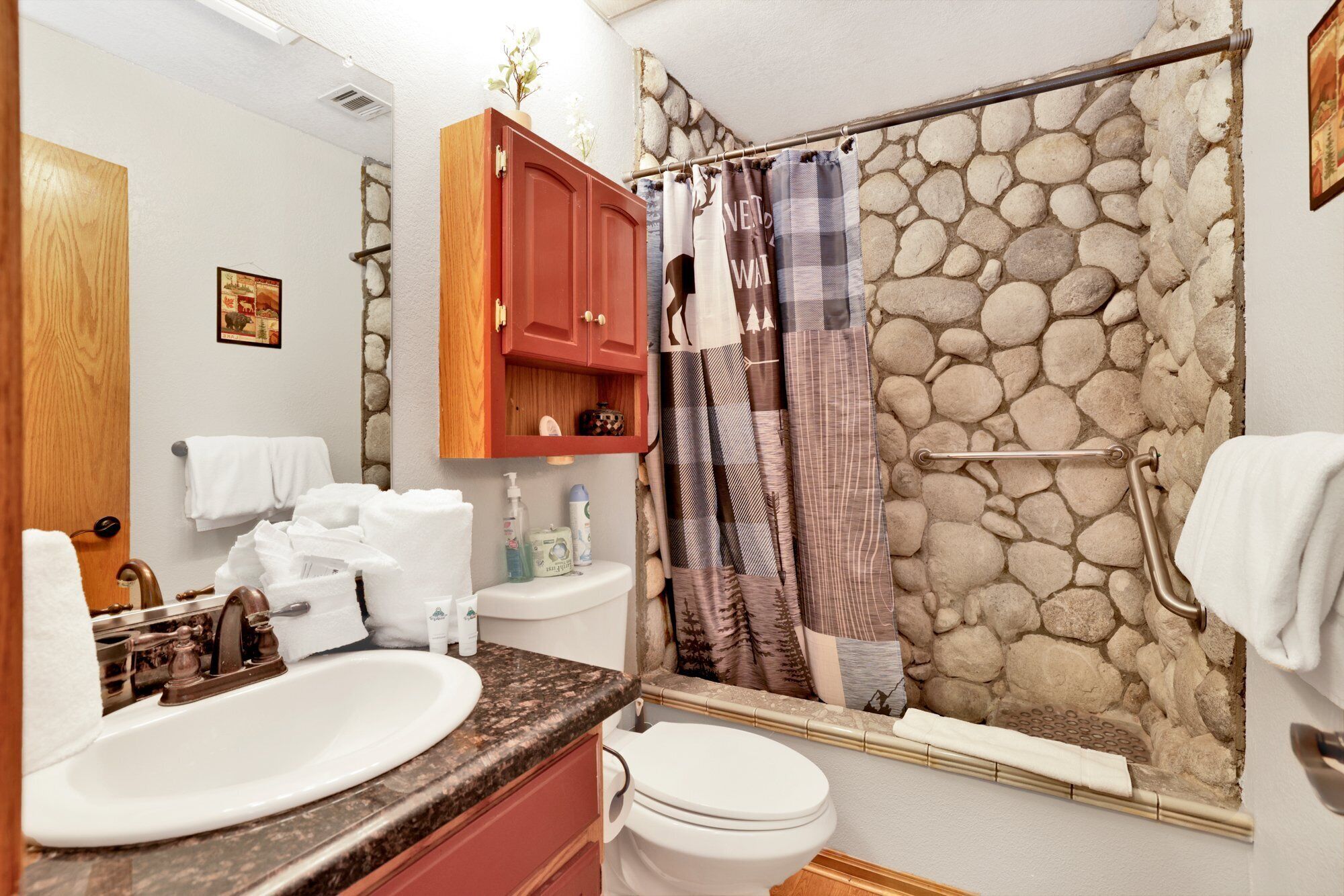 A bathroom with a stone shower wall, a wooden cabinet, a sink, and a toilet.