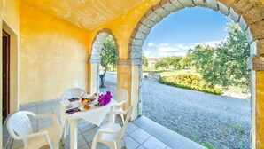 Un coin repas avec une table dressée pour deux personnes, des chaises blanches et une vue sur un jardin, accessible par une arche.