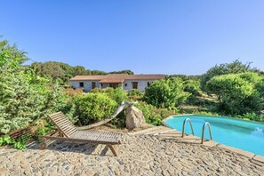 A stone pool with lounge chairs, a stone pathway, and a stone building with a tiled roof in a lush, green setting.