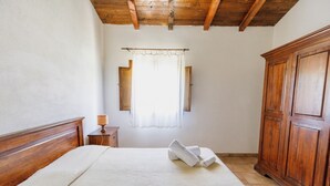Una camera da letto con testiera in legno, un letto con copripiumino bianco e testurizzato, un comodino con lampada, una finestra con tende e un armadio in legno.
