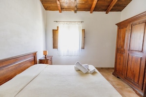 Une chambre à coucher avec une tête de lit en bois, un lit recouvert d’une courtepointe blanche à texture, une table de chevet avec une lampe, une fenêtre avec des rideaux et une armoire en bois.