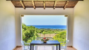 Un balcone con tavolo e sedie, affaccia su una vista panoramica del mare e del verde.