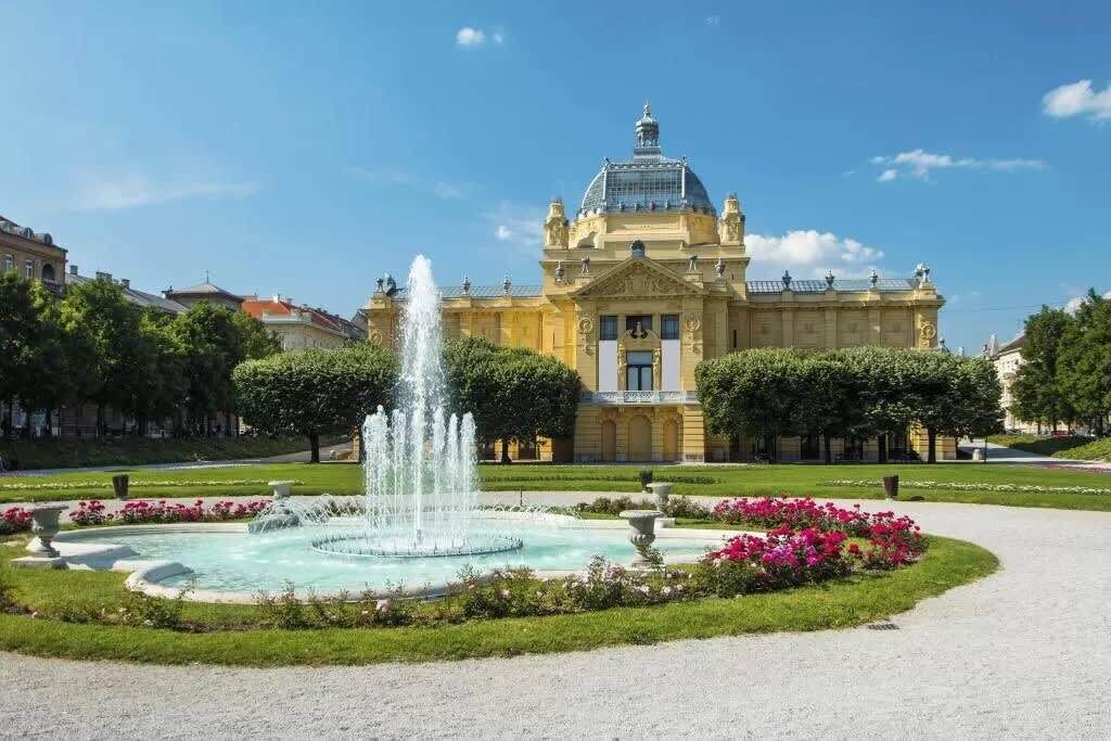 A grand building with a fountain in the center, surrounded by well-maintained gardens and a clear sky.