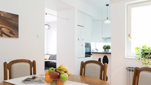 A dining area with a wooden table, chairs, and a fruit bowl. A kitchen is visible in the background.
