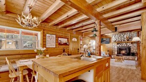 A rustic cabin interior with a wooden dining area, a stone fireplace, and a large window overlooking a forested area. - Binky's Mountain Hideaway #2455 (Big Bear Lake)