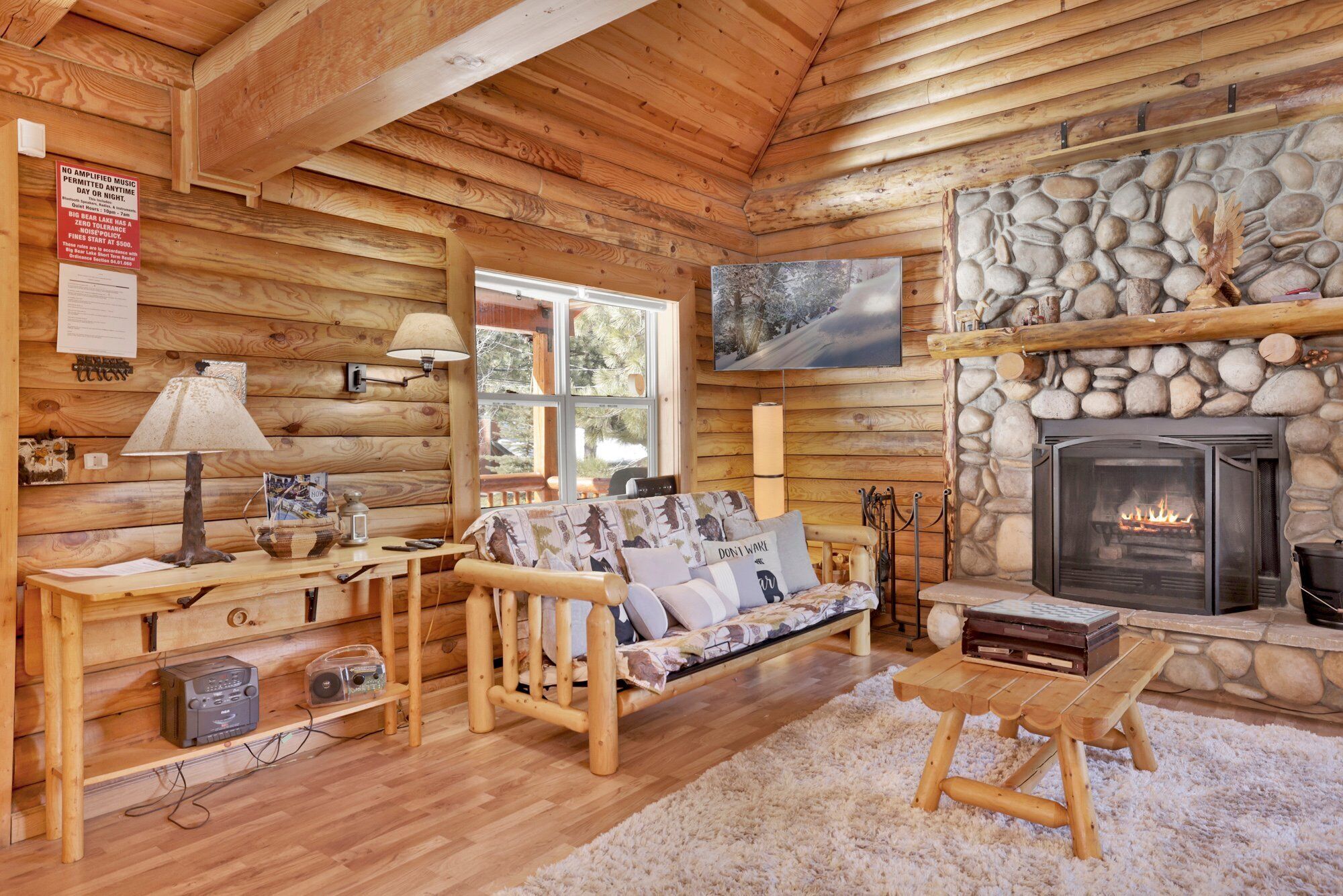 A cozy cabin interior with a stone fireplace, wooden furniture, and a television mounted on the wall.