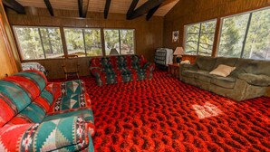 A living room with a red patterned carpet, two sofas with colorful upholstery, and large windows offering a view of trees. - Makin' Memories AT Summit #1683 (Big Bear Lake)