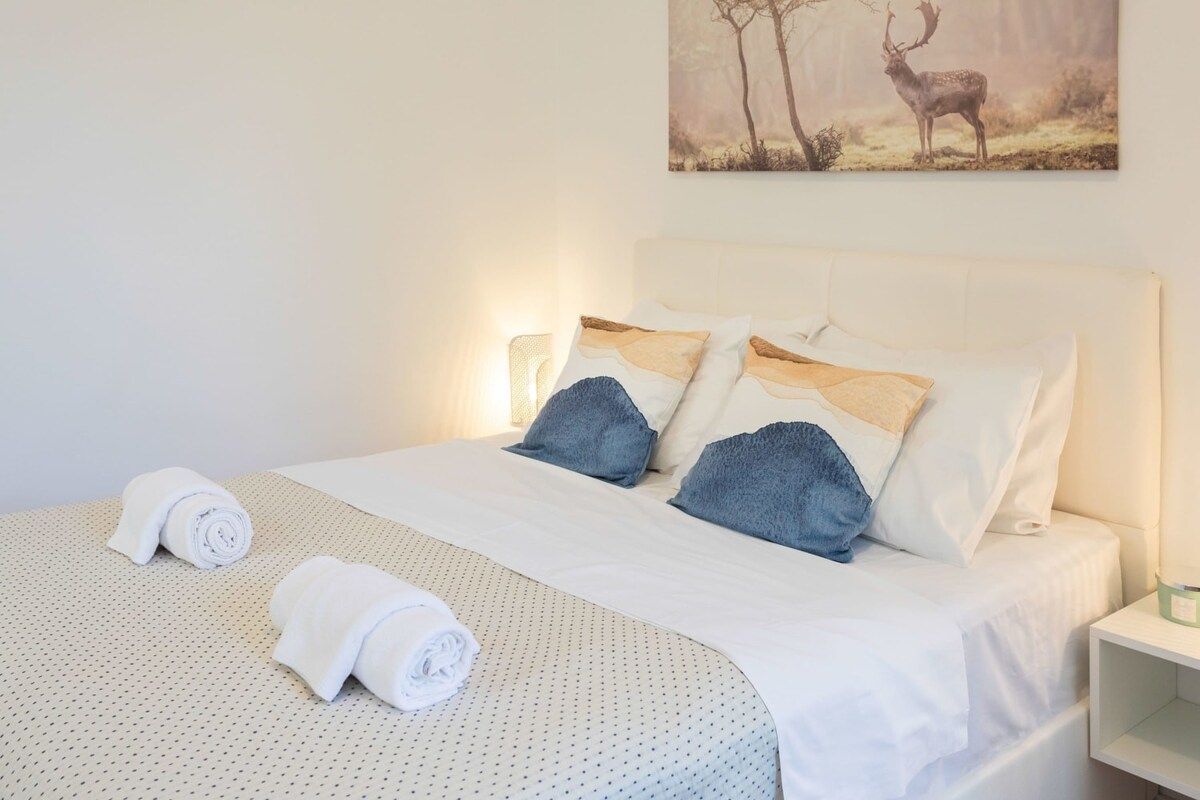 Un lit bien fait, avec du linge de lit blanc et deux serviettes enroulées, un tableau représentant des cerfs accroché au mur, et une table de chevet avec une lampe.