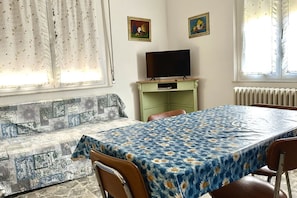 Une pièce comprenant un canapé, une table recouverte d’une nappe à motifs floraux, un téléviseur posé sur un meuble et deux tableaux encadrés accrochés au mur.