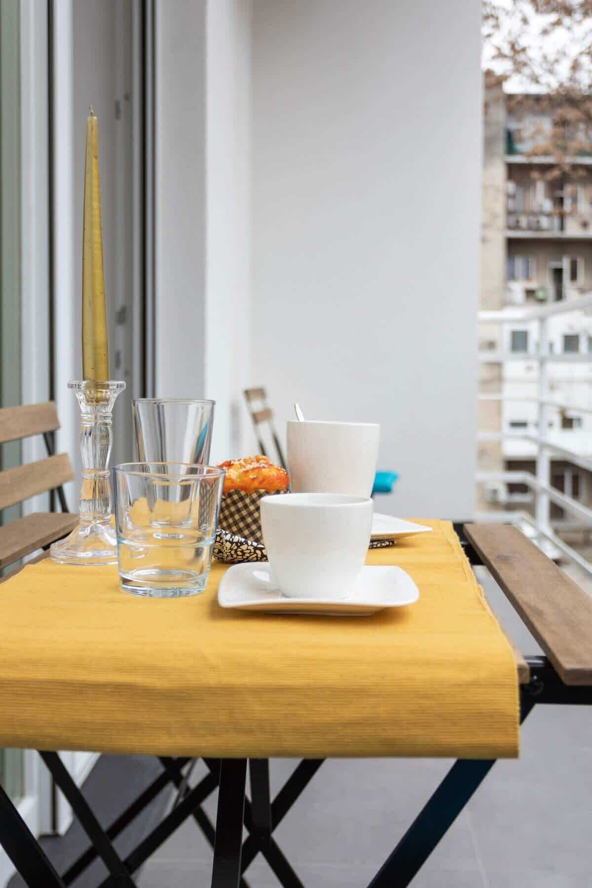 Una mesa de madera con vasos transparentes, una taza blanca y una vela en un balcón con vistas a edificios.