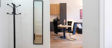 A modern interior with a coat rack, a round dining table with chairs, a kitchen area, and a large mirror on the wall.