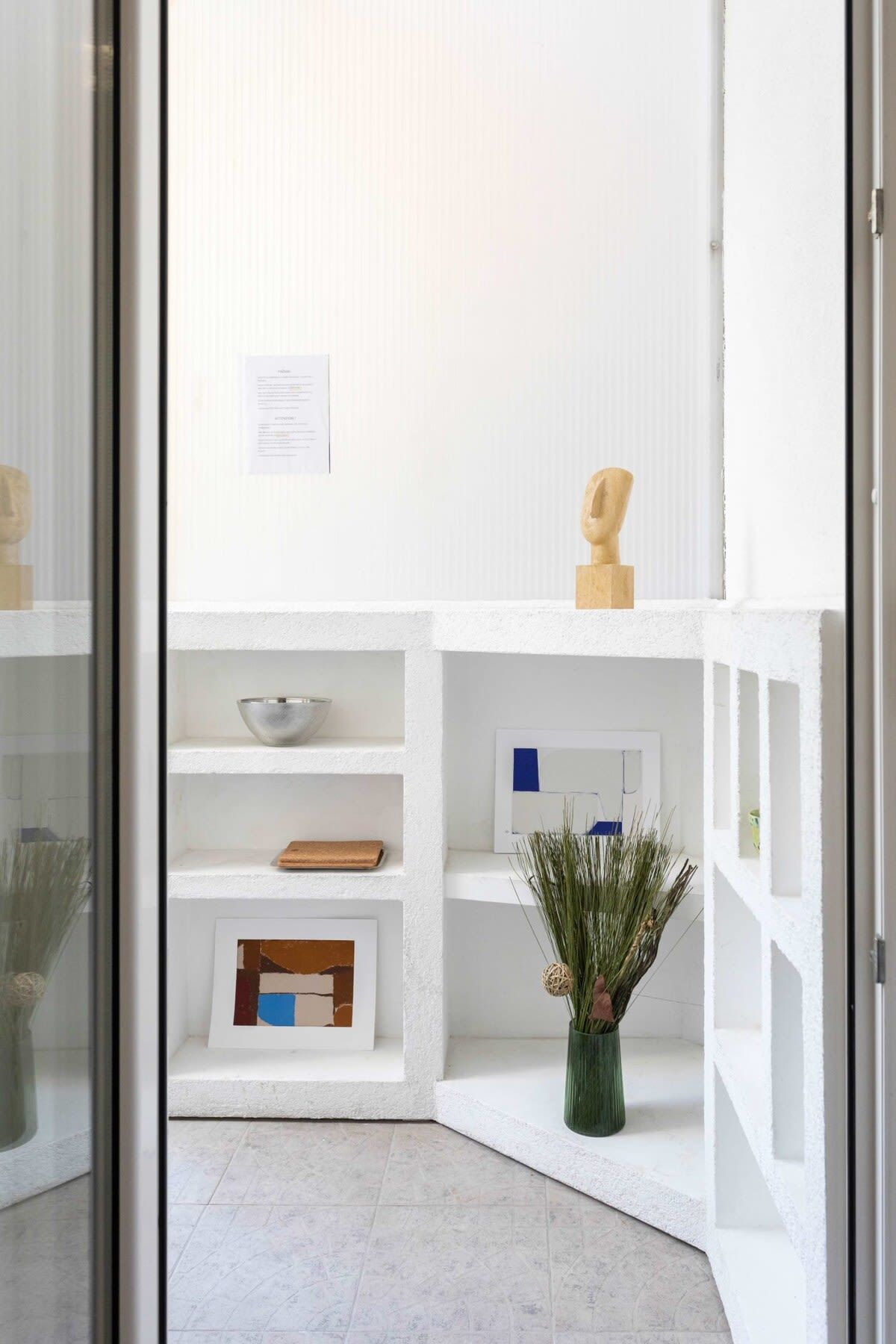 Een modern interieur met witte open planken waarop diverse objecten staan, een ingelijste afbeelding en een vaas met gedroogde planten.