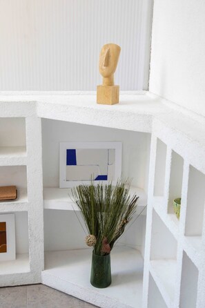 Ein heller, moderner Innenraum mit einer geometrischen Skulptur, einem gerahmten abstrakten Kunstwerk und einer grünen Vase mit Trockenpflanzen.