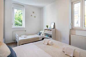 A bedroom with two single beds, a window, a radiator, and a small shelf with a plant and picture frame.