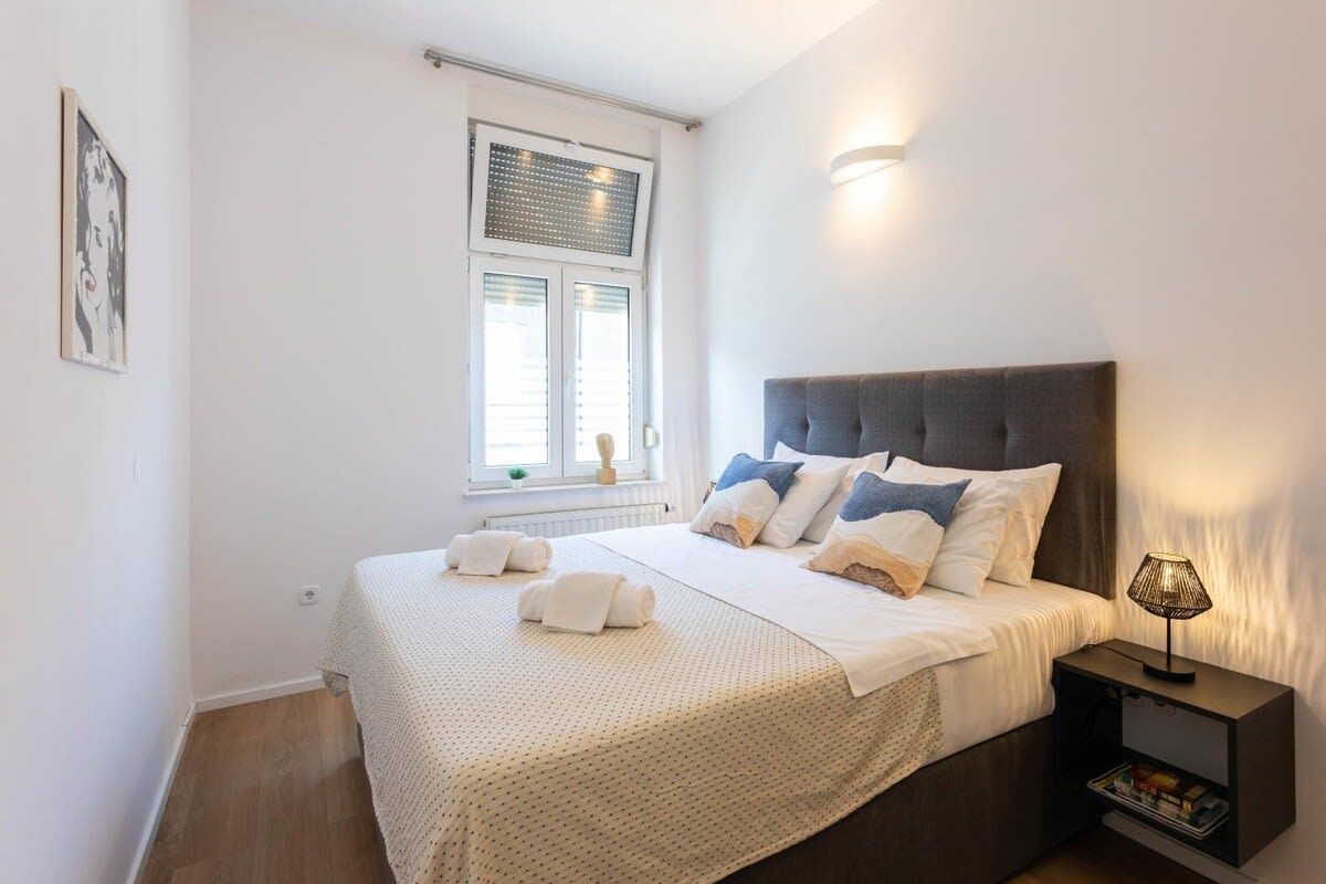 A bedroom with a bed, pillows, a nightstand, a lamp, a window with blinds, and a wall-mounted light.