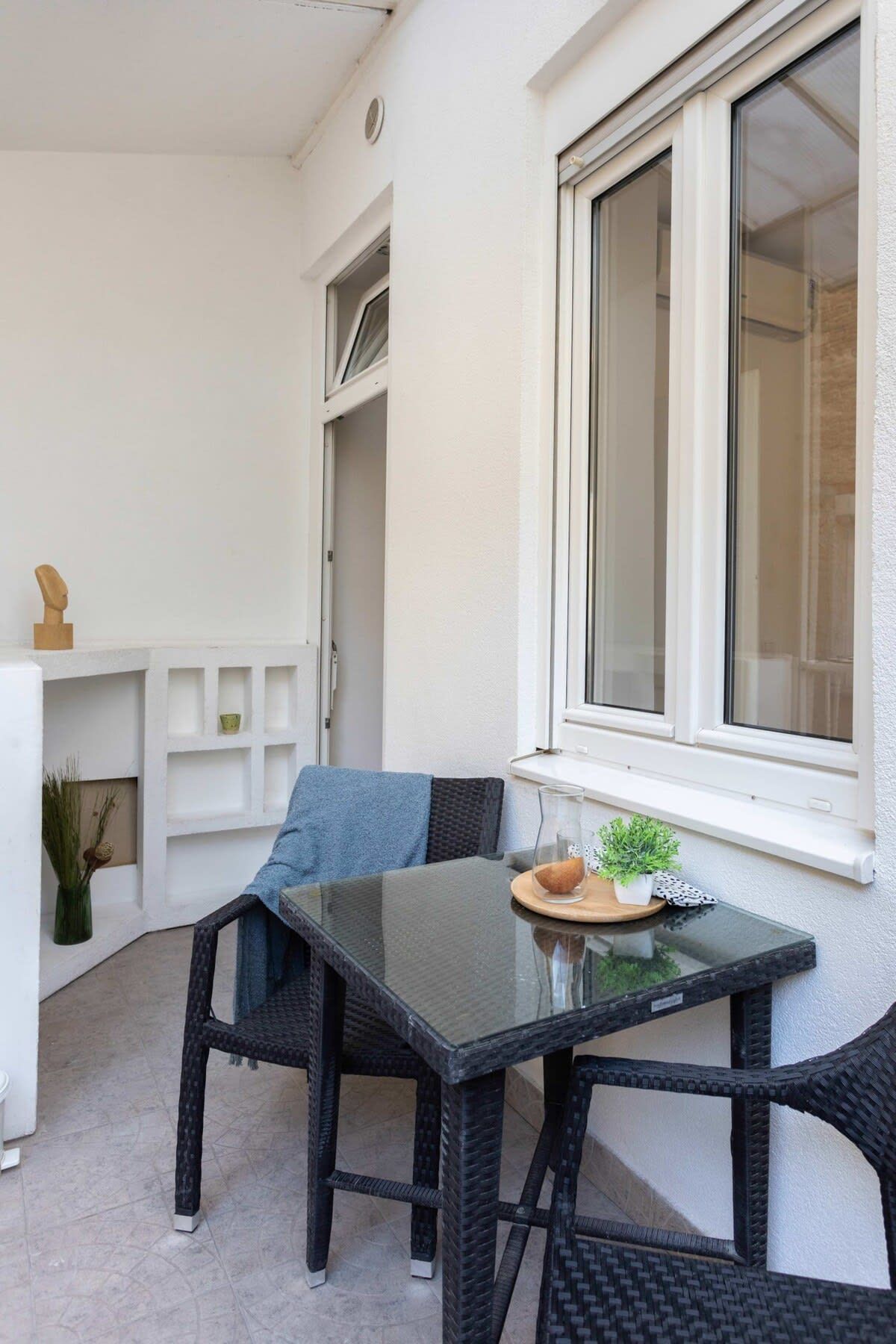 Una pequeña zona de estar al aire libre con una mesa de vidrio y sillas de mimbre, una ventana con persianas y una pared blanca con un estante.