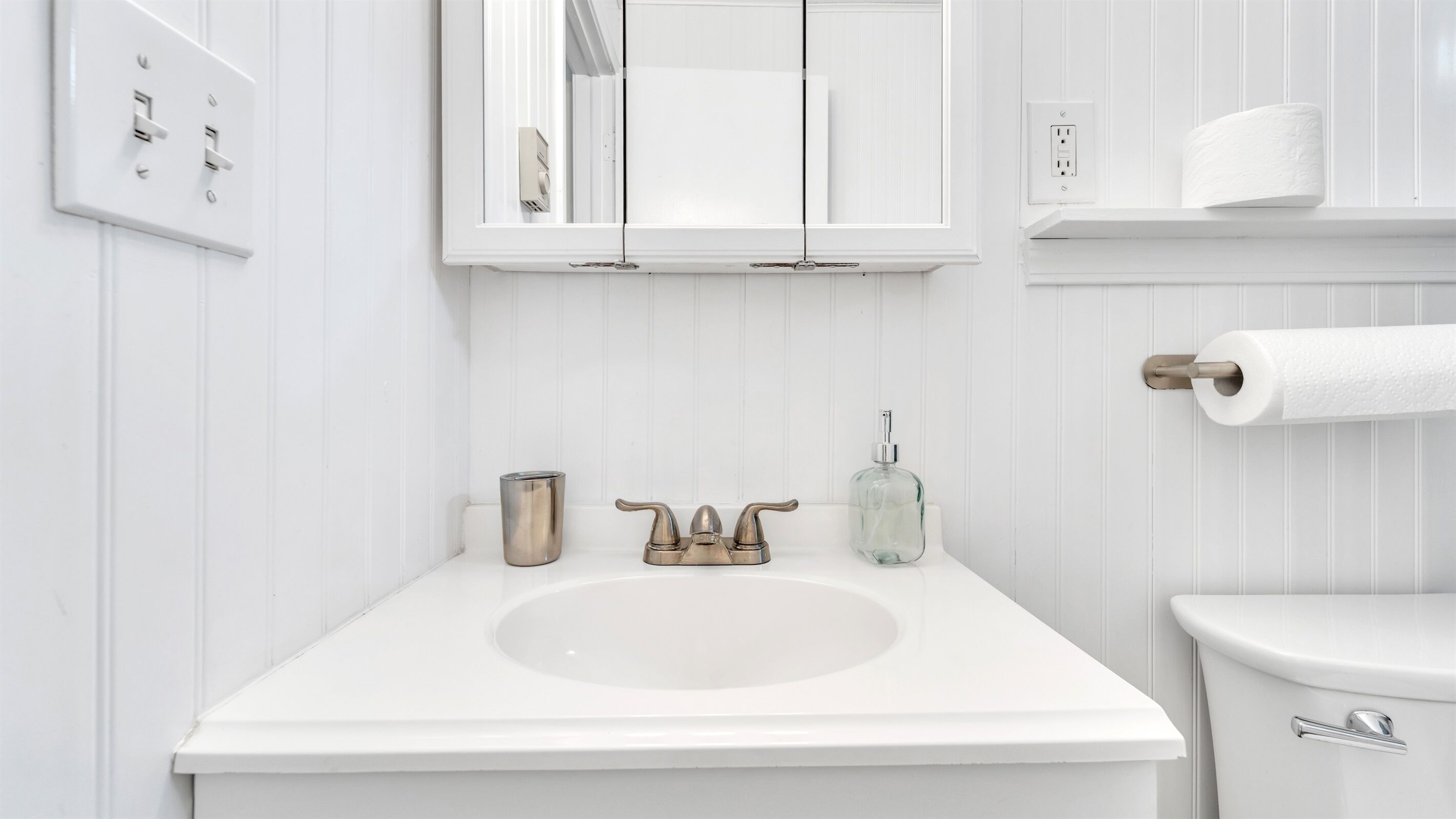A white bathroom with a sink, a mirror, a toilet, and a roll of toilet paper on a shelf.