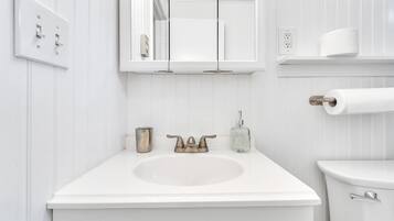 A white bathroom with a sink, a mirror, a toilet, and a roll of toilet paper on a shelf.