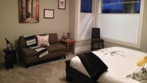 Un dormitorio con una cama grande, un sofá marrón, una mesita y una silla. Hay una ventana con persianas y un marco con un cuadro en la pared.