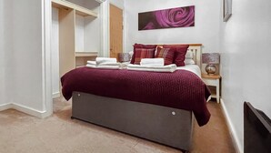 A neatly made bed with a grey headboard, a red blanket, and several pillows. There is a wooden nightstand with a lamp and a decorative item beside the bed. A large painting hangs on the wall above the bed.