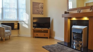 A modern living room with a fireplace, a flat-screen TV on a wooden stand, a plaid armchair, and a decorative wall piece.