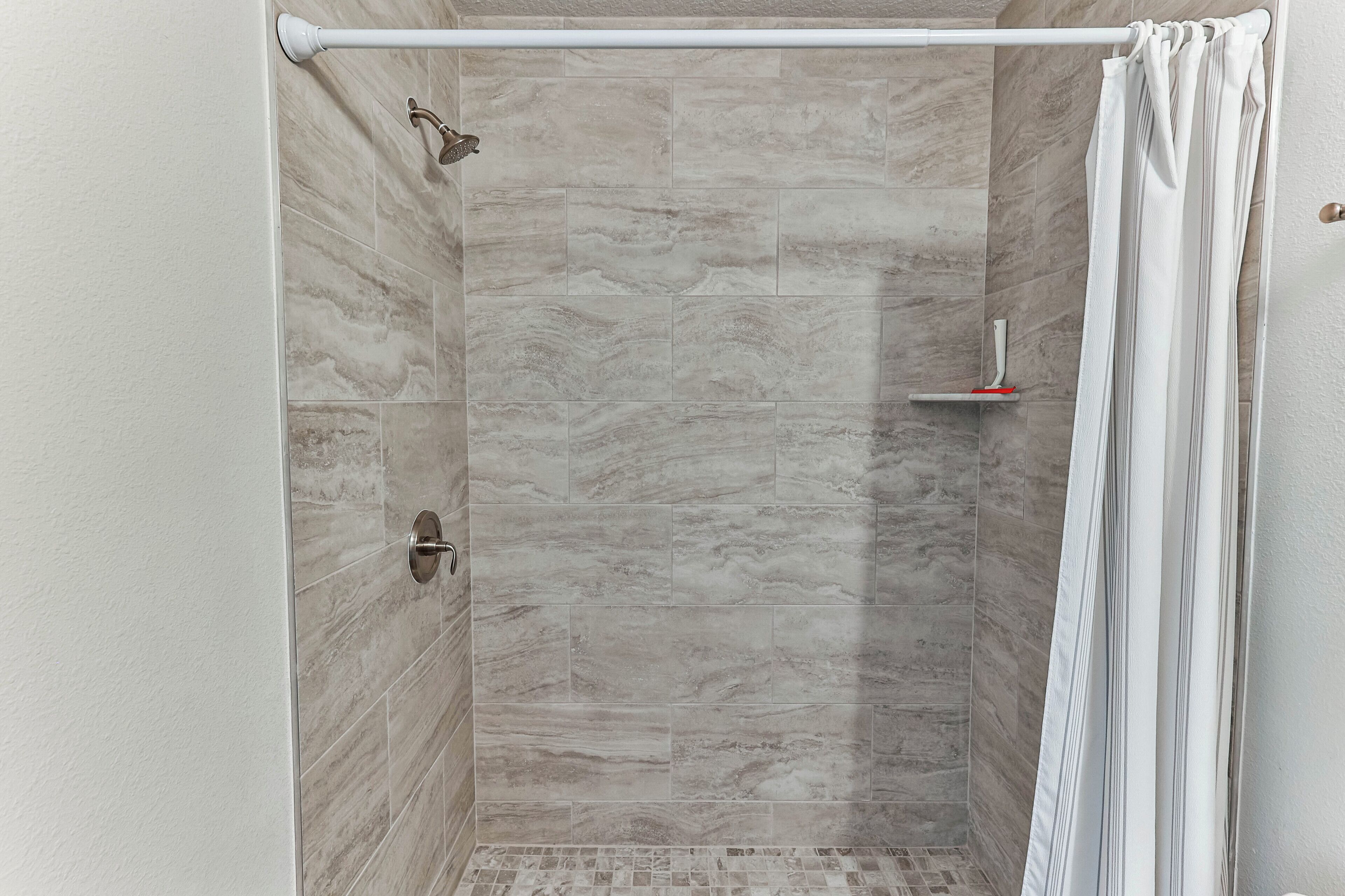A shower with a white curtain, a handheld showerhead, and a small shelf with a red object.