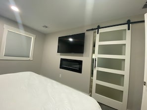 Un dormitorio con cama, televisor montado en la pared, armario con puerta corredera de vidrio y ventana con persianas.
