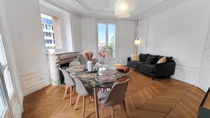 A dining area with a glass table, white chairs, and a fireplace. There is a sofa and a window with a view of buildings. - Amazing refurbished flat in Paris V (Paris)