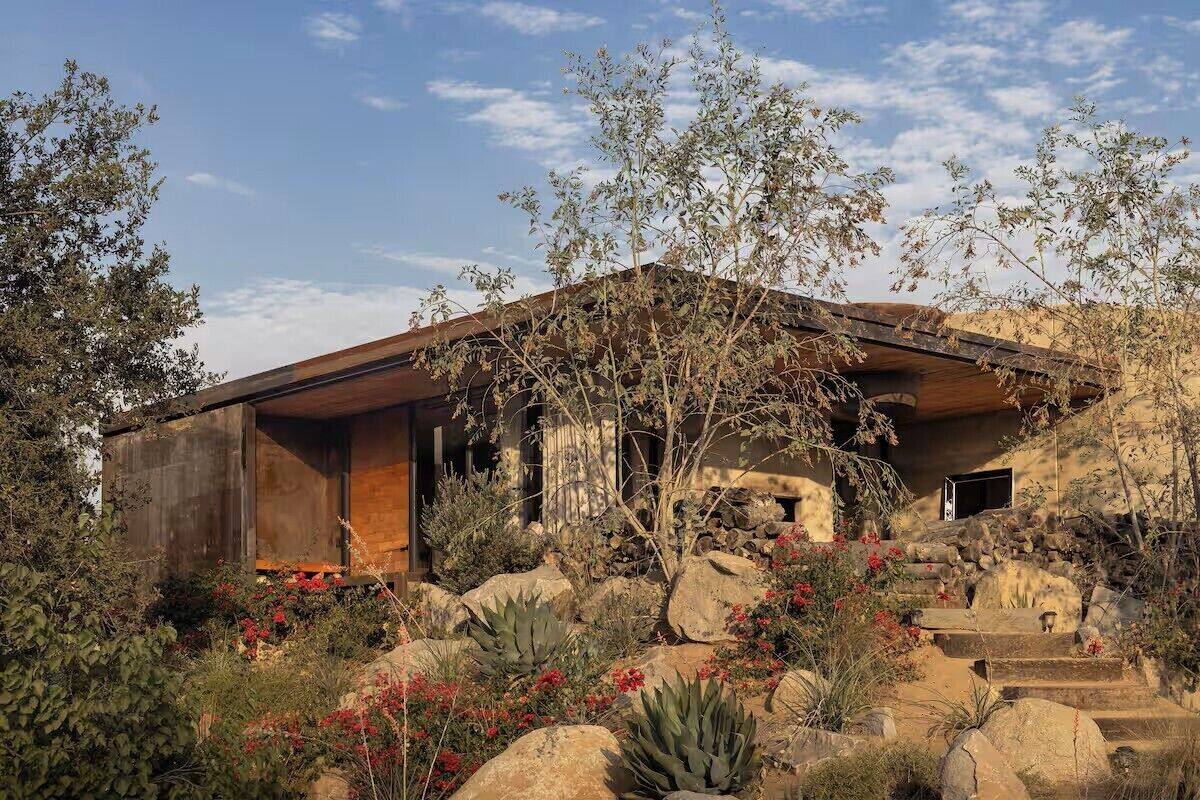 A modern house with a stone facade, wooden beams, and a landscaped garden with rocks and cacti.