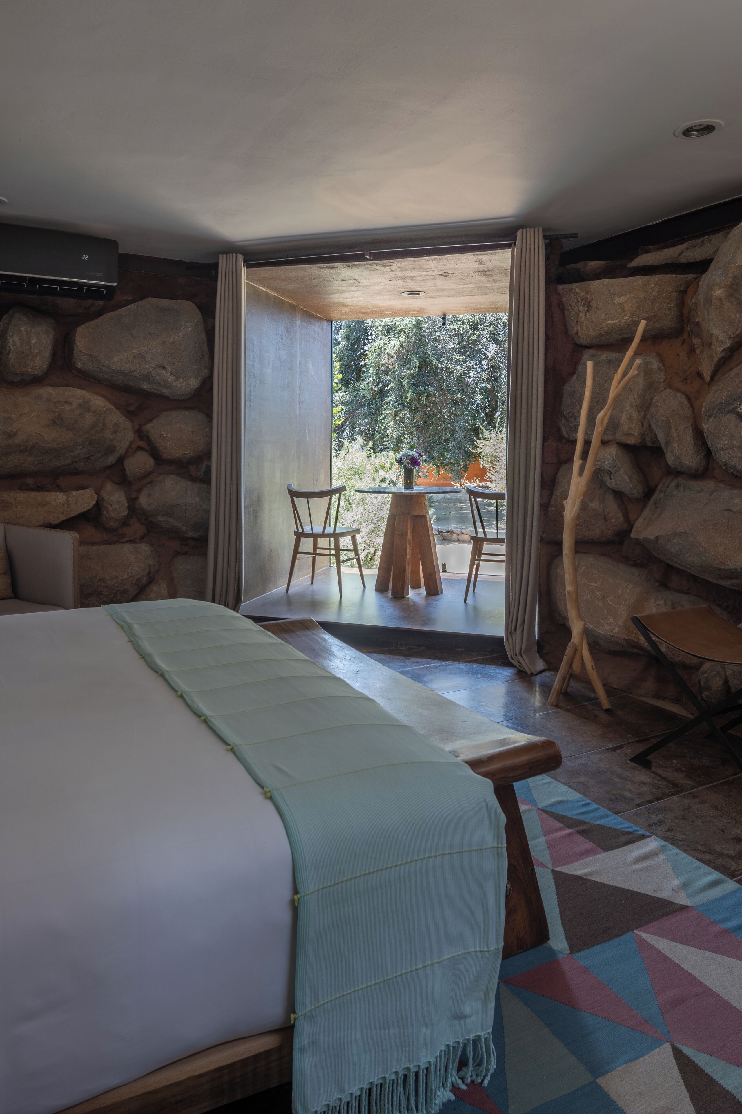 A bedroom with a large bed, a wooden chair, a small table with a vase, and a view of the outdoors through a glass door.