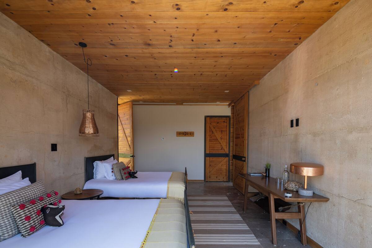 A room with two beds, a wooden ceiling, a small table with a lamp, and a door leading to another room.