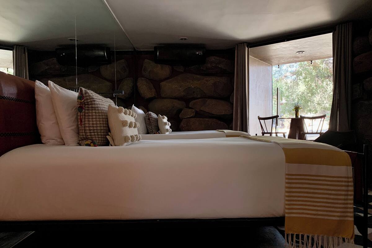 A large bed with multiple pillows, a stone wall, and a view of the outdoors through a window.